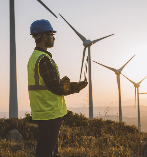 Ingenieur mit Schutzhelm und Warnweste inspiziert einen Windpark bei Sonnenuntergang