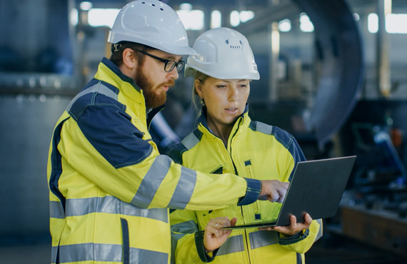 Zwei Industriearbeiter in Schutzhelmen und Warnschutzjacken schauen gemeinsam auf einen Laptop in einer Fabrikhalle
