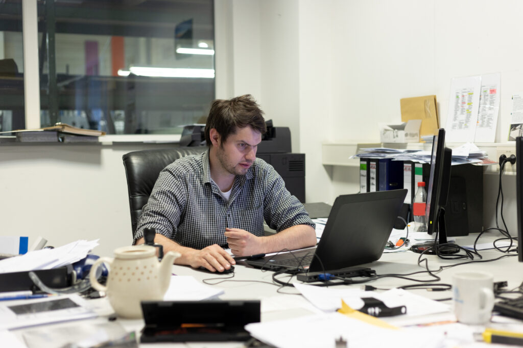 Mann arbeitet konzentriert an einem Laptop an einem unordentlichen Büroschreibtisch