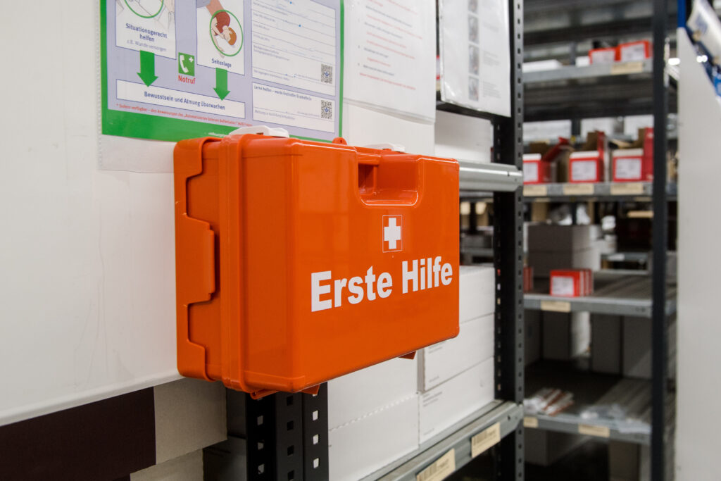 Orangefarbener Erste-Hilfe-Koffer an einem Regal in einem Lager, daneben ein Erste-Hilfe-Aushang an der Wand