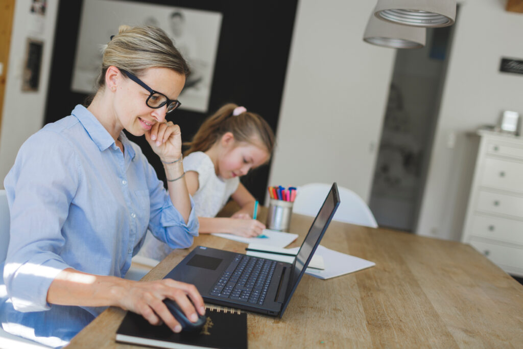 Frau arbeitet im Homeoffice am Laptop, während ein Kind neben ihr am Tisch malt